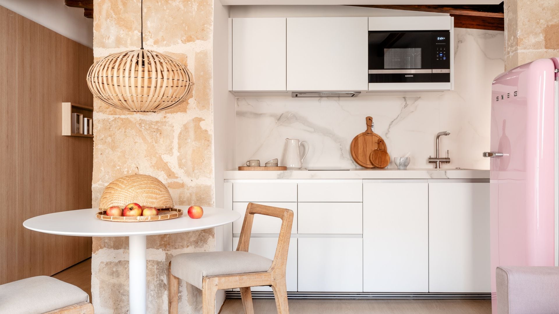 Cocina con comedor, columnas de piedra, cocina blanca, nevera Smeg de color rosa, mesa redonda, lámpara de techo de fibras vegetales 