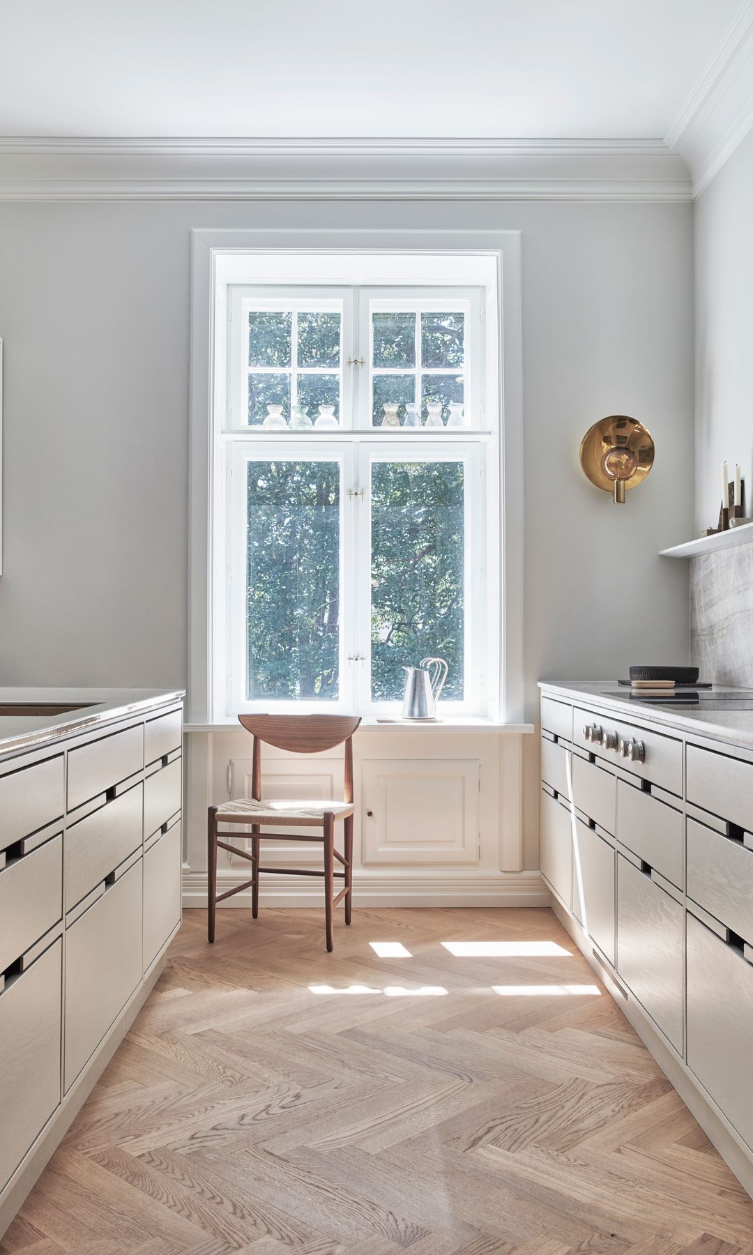 Cocina blanca distribución en paralelo con suelo de madera, ventana con perfiles blancos y silla de madera